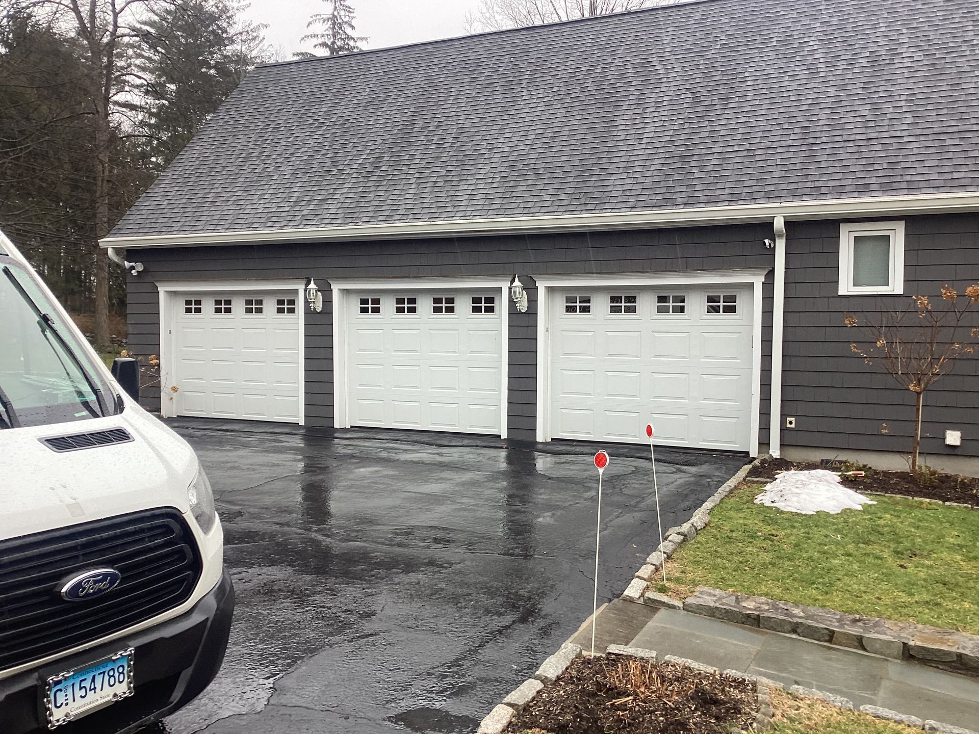 White garage with three doors beside a house, viewed across a wet driveway on a cloudy day
