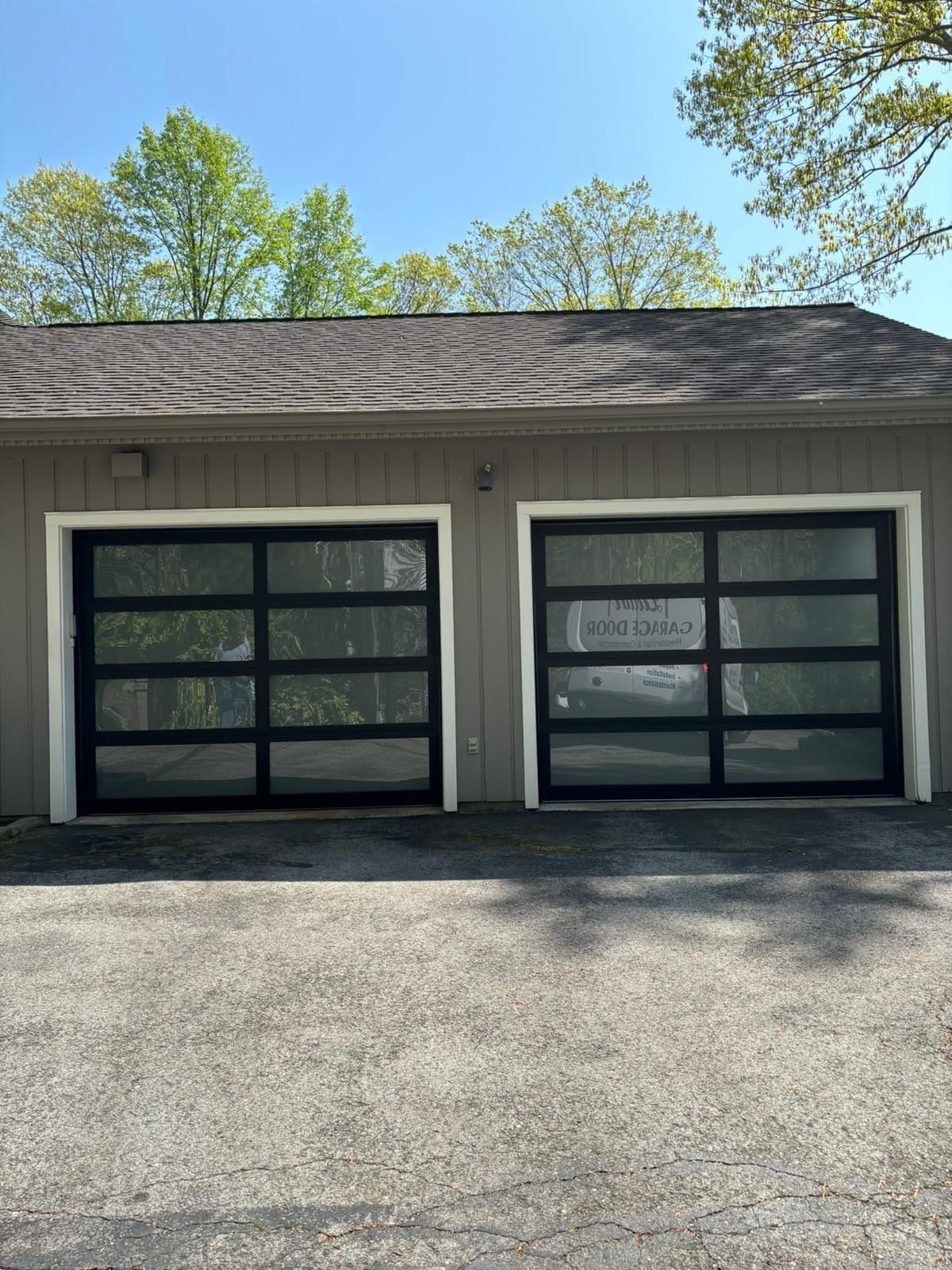 Two black-framed garage doors on a white building with a gray shingled roof and trees in back