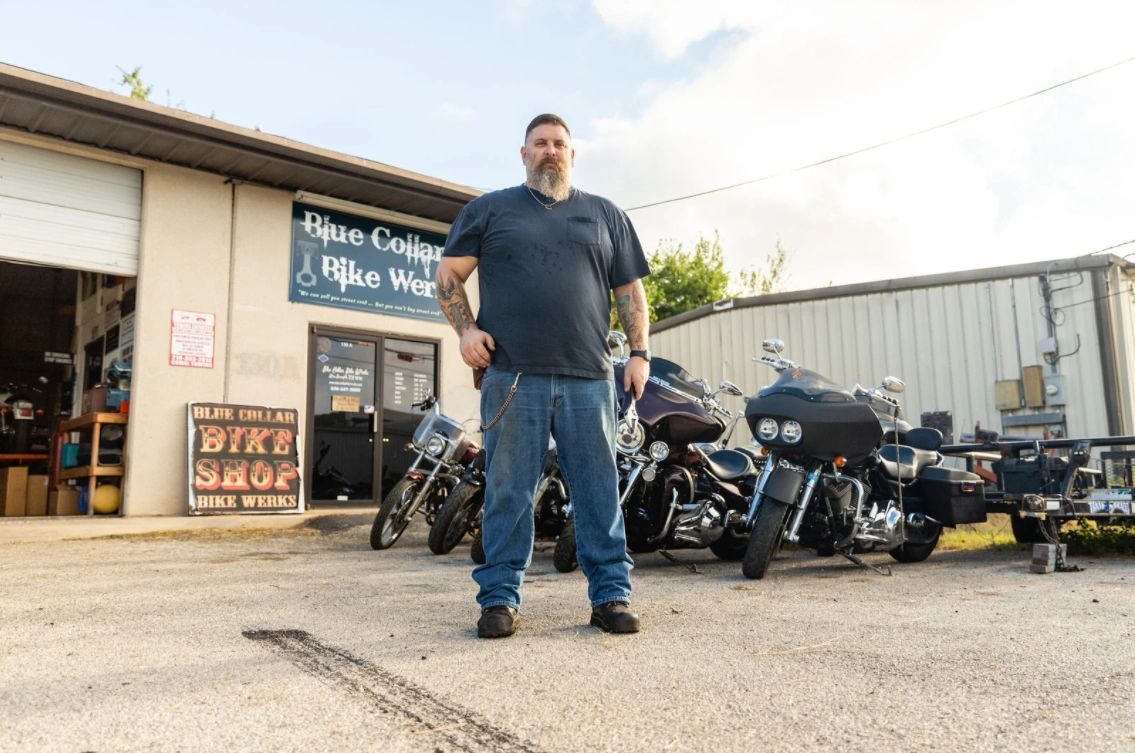 Man in front of bike shop with motorcycles. He's wearing a dark shirt and jeans.