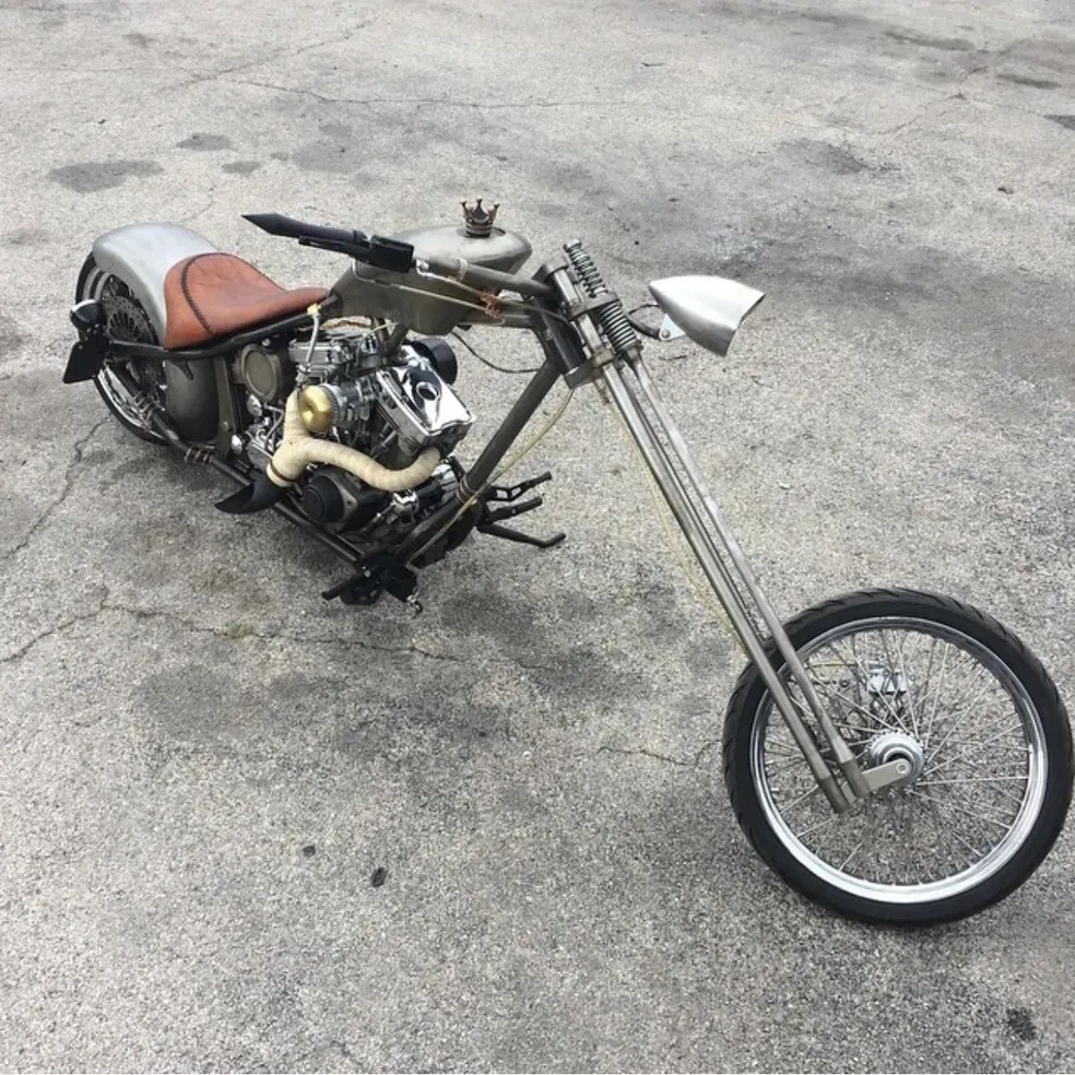Custom chopper motorcycle on a concrete surface, metallic frame, brown seat, elongated front fork, and chrome headlight.