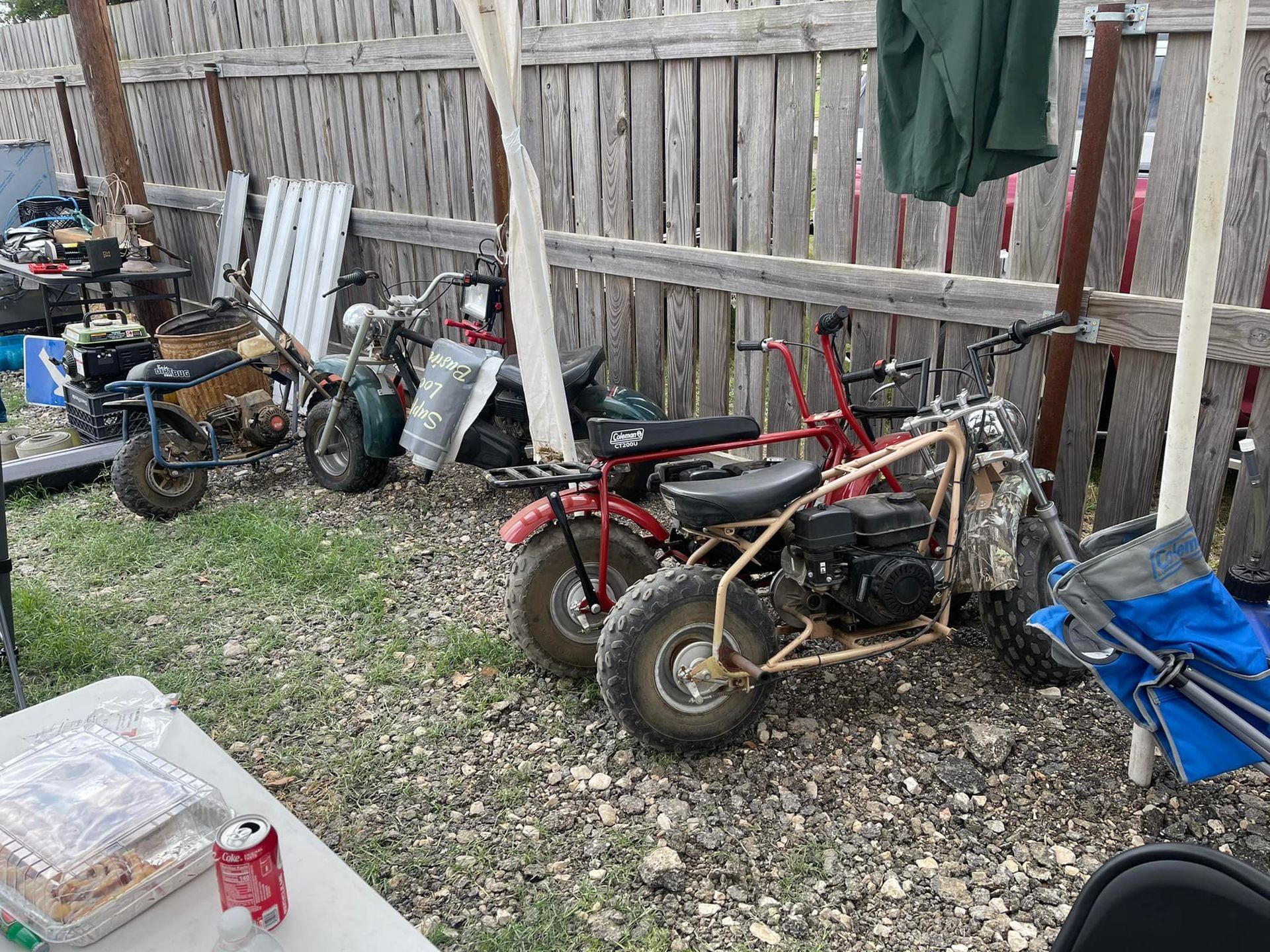 Mini bikes lined up along a wooden fence at an outdoor gathering.