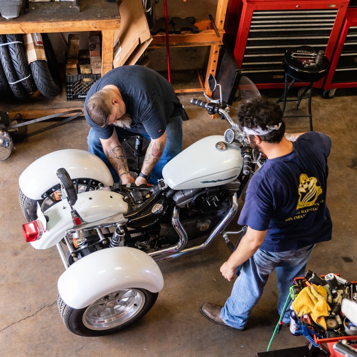 Two men working on a white trike motorcycle in a garage, one man is kneeling, the other is standing.