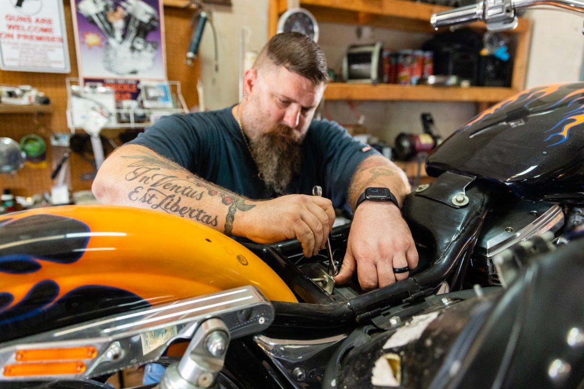 Man with beard working on a motorcycle in a garage. He's wearing a black shirt, has tattoos and a watch. Motorcycle has orange flames.
