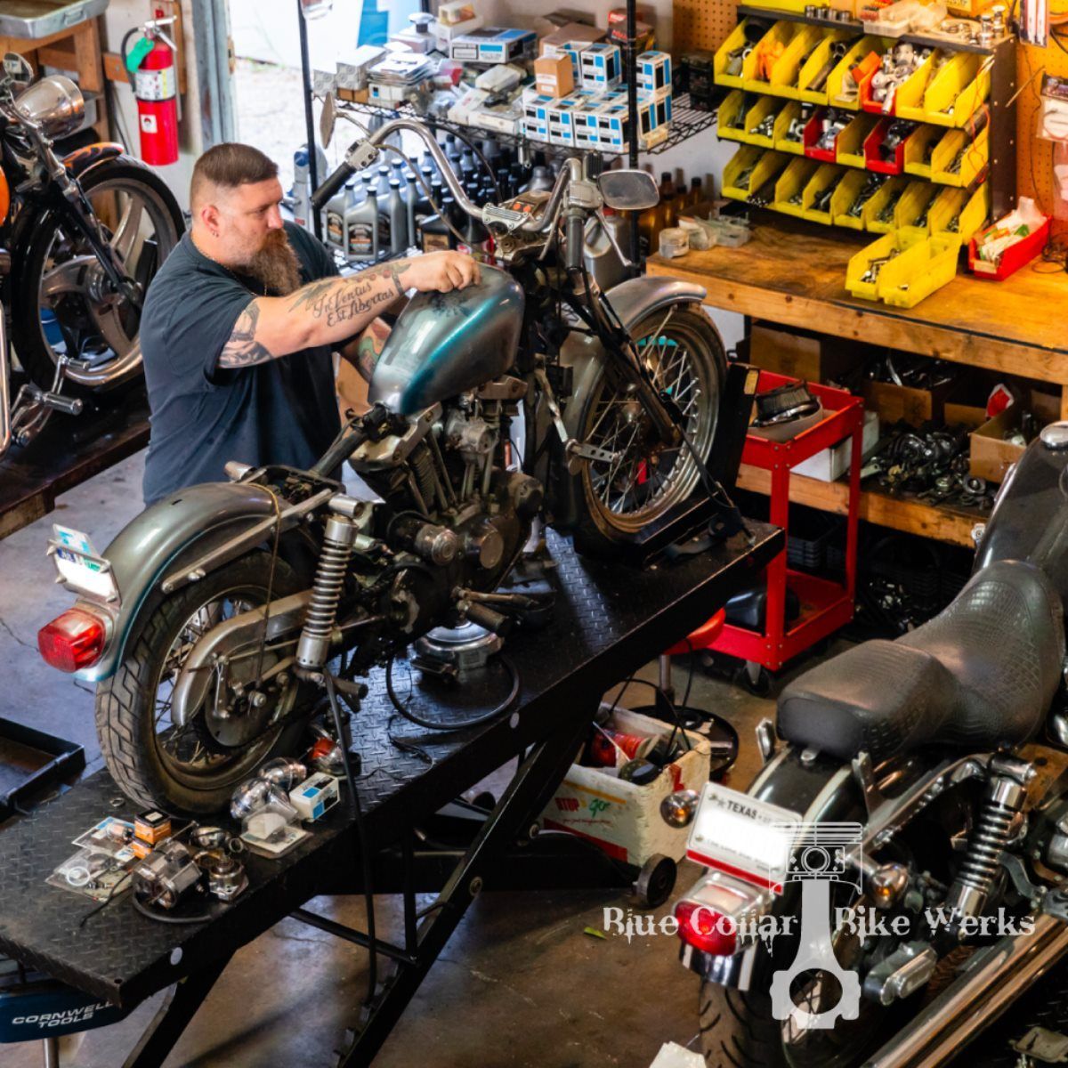 A man with tattoos works on a motorcycle in a well-lit workshop. Tools are visible.