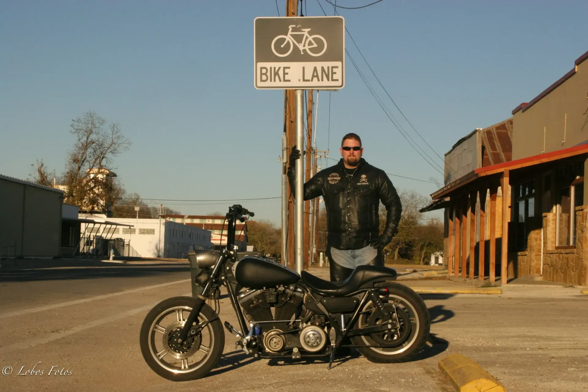 Man in leather jacket stands next to motorcycle under a bike lane sign on a street.