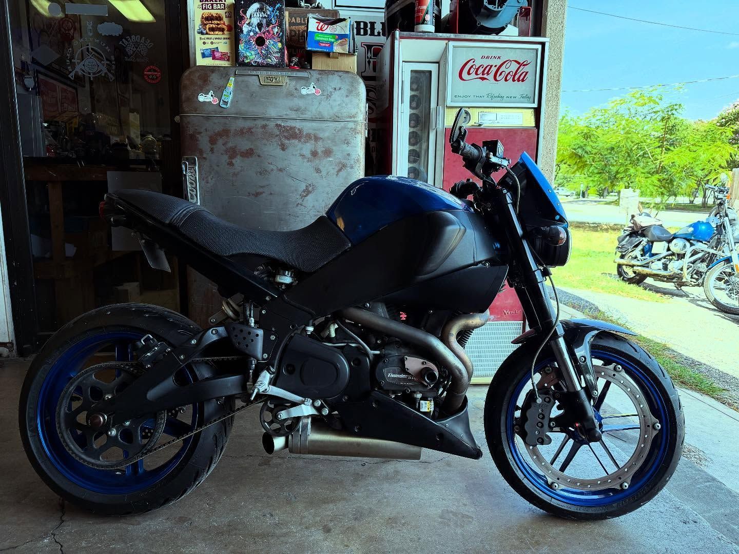 Blue and black motorcycle parked inside a garage, with a vintage Coca-Cola machine in the background.