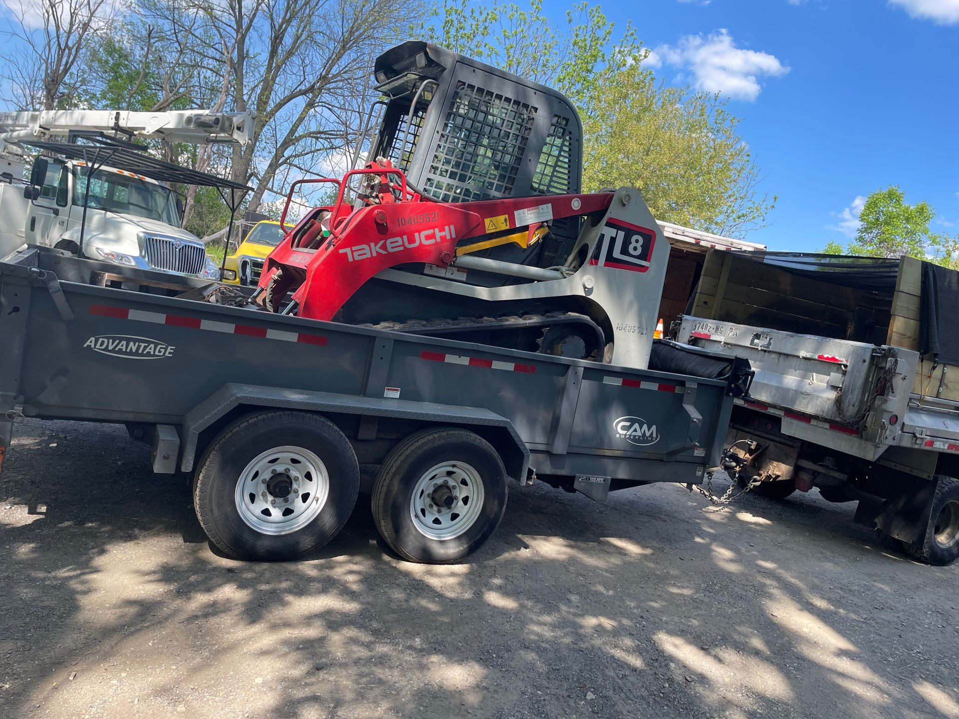 Red and silver skid steer on a trailer, parked next to a dump truck, outdoors on a sunny day.