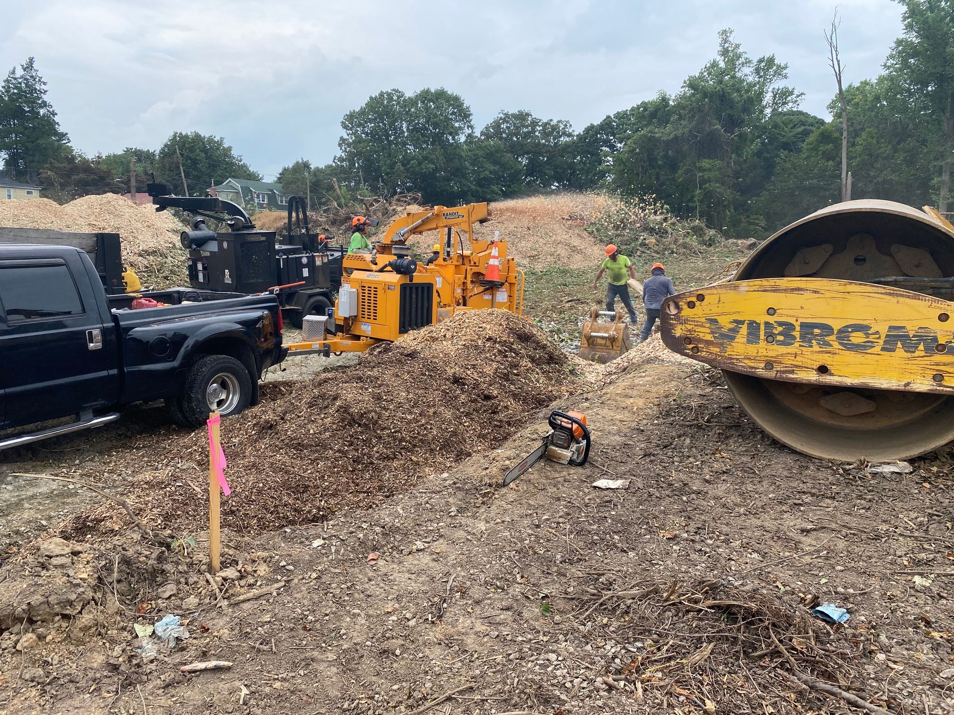 Workers using a wood chipper, truck, and roller on a pile of wood chips outdoors.