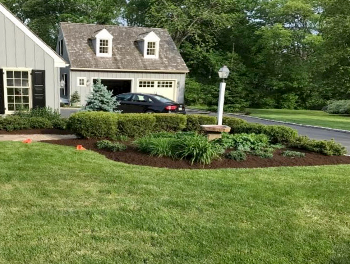 A suburban home with a gray garage, a manicured lawn, a dark car, and a landscaped garden bed with a central lamppost.