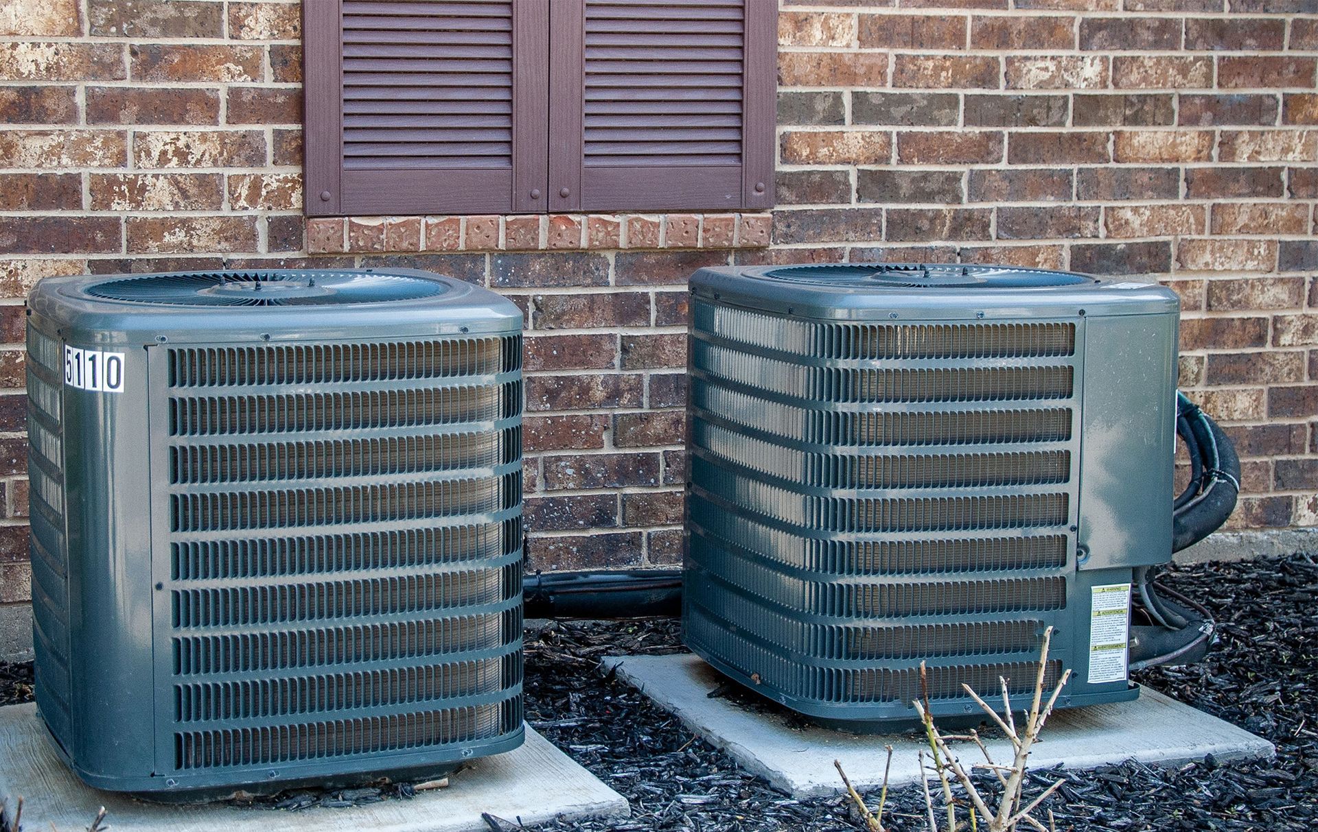 Two outdoor HVAC units beside a brick wall and window shutters