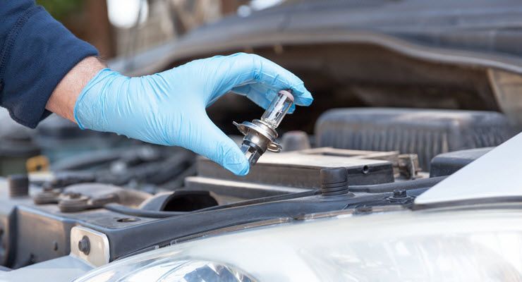 Hand in blue glove holding car headlight bulb near open car hood.