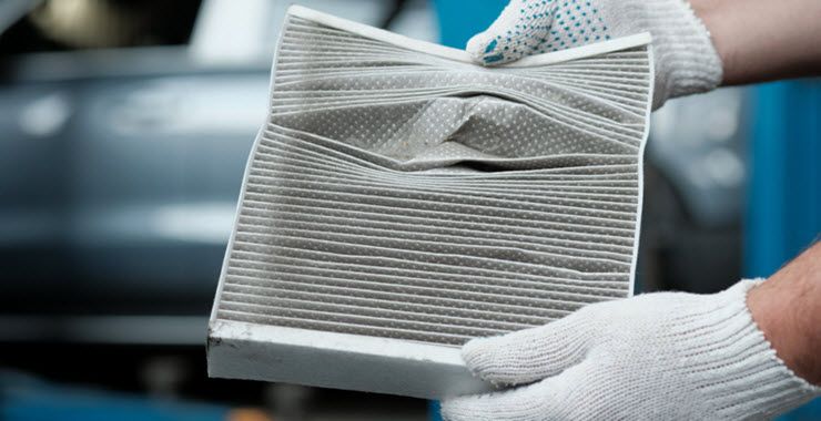Gloved hands holding a dirty, compressed car cabin air filter, showing wear and damage.