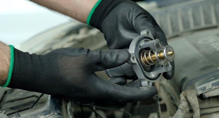 Hands in black gloves holding a car thermostat against a blurred engine background.