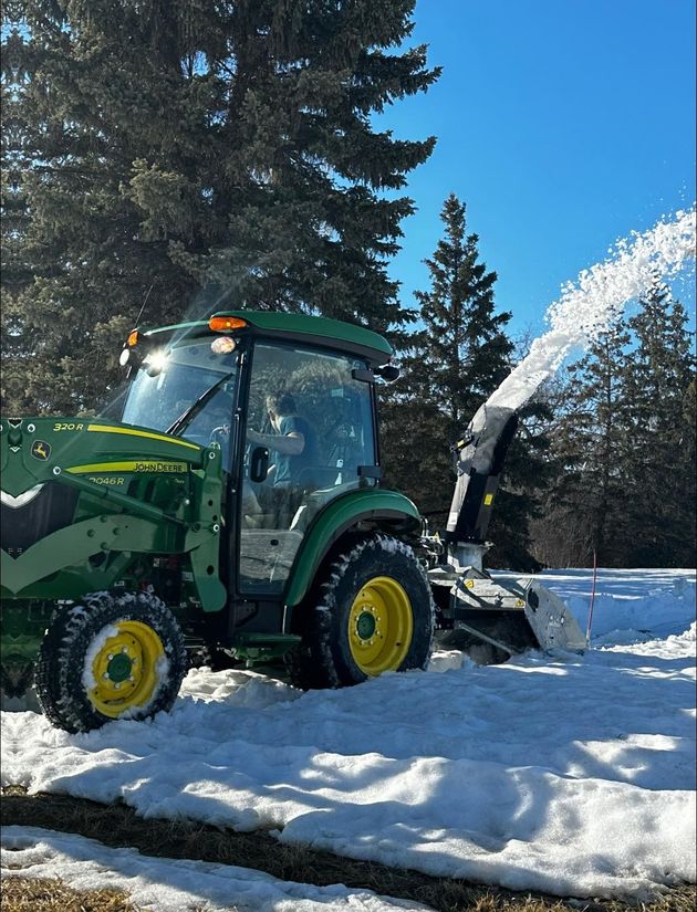 A green John Deere tractor with a rear-mounted snow blower throwing snow into the air on a sunny, winter day.