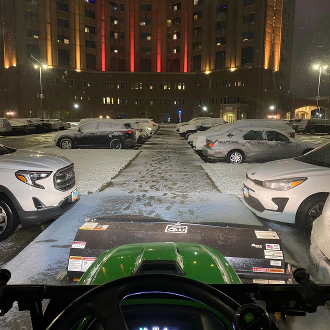 Snowy parking lot at night, viewed from a green vehicle facing a lit building with parked cars.