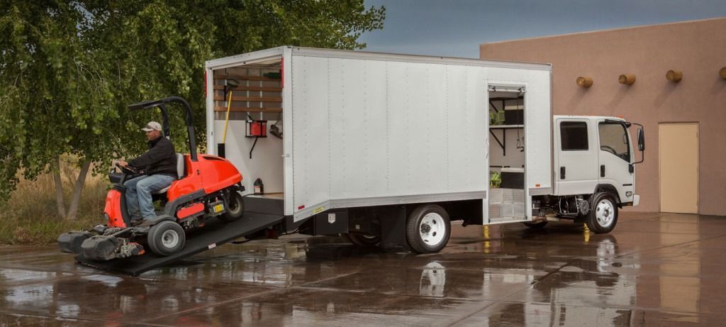 Man on a small orange tractor drives onto a truck ramp. Truck is next to a tan building, with water on the ground.