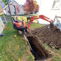 A small excavator is digging a hole in the ground in front of a house.