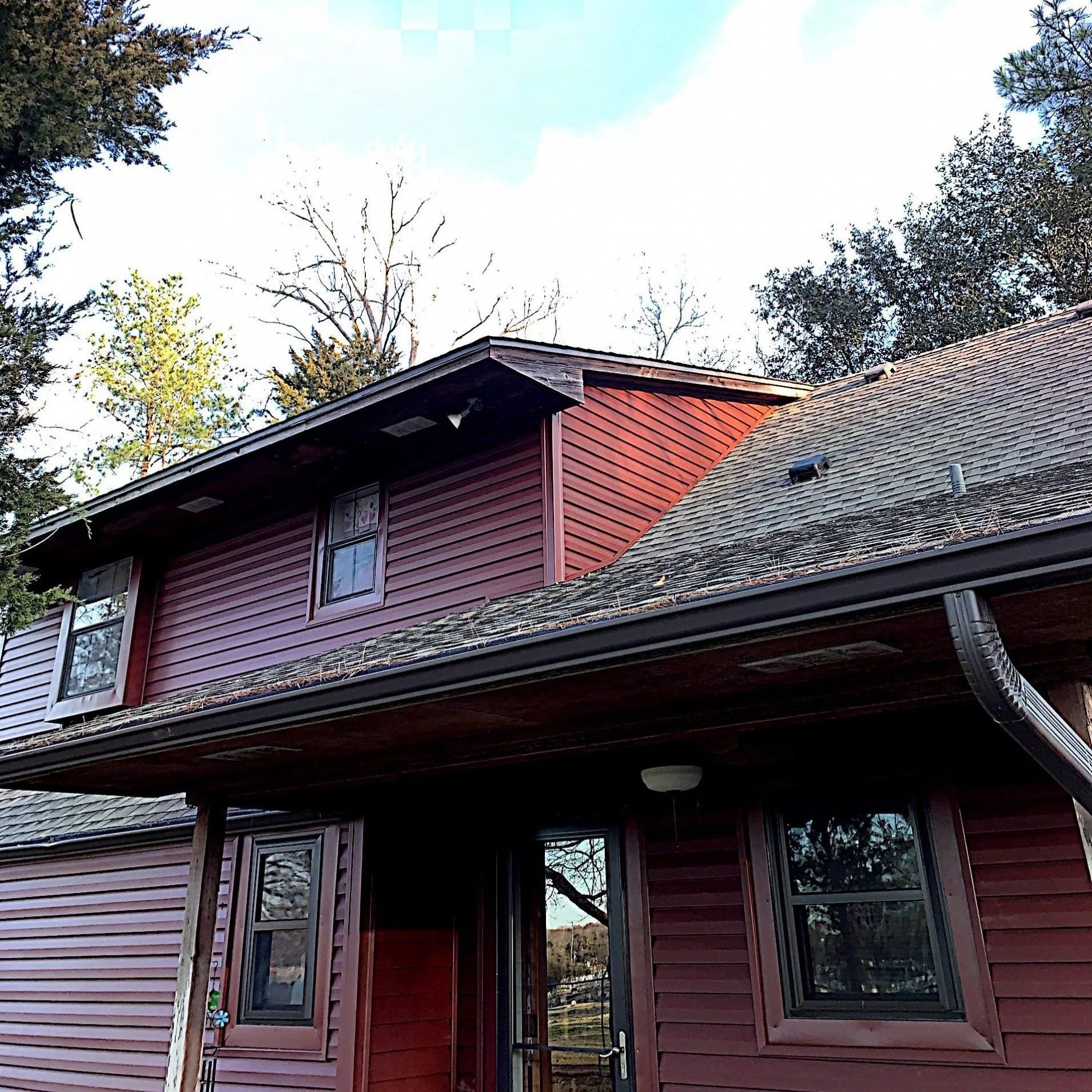 Red wooden house with a gray shingled roof and covered porch, seen from below among trees