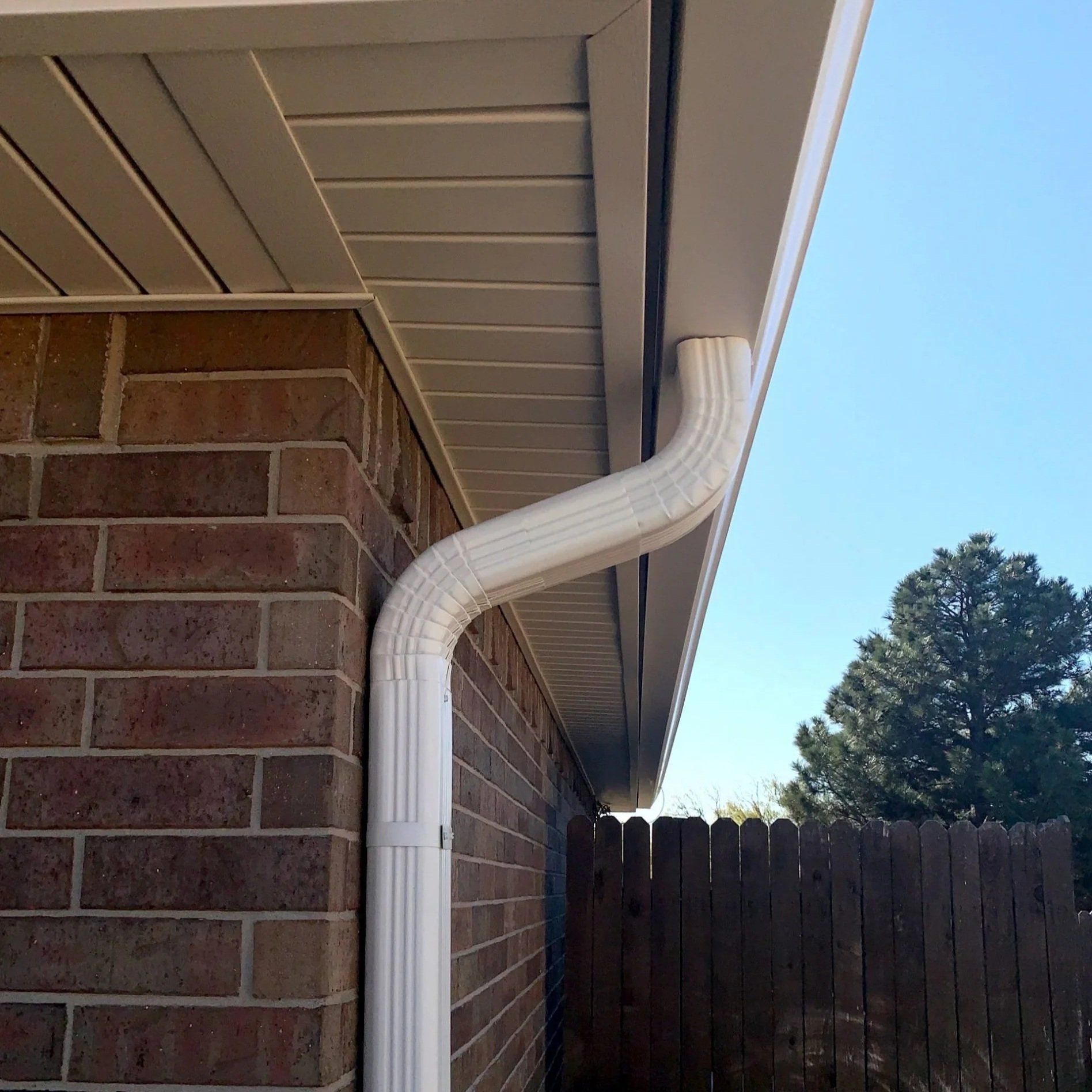 White downspout running along a brick house corner under the roof eave, beside a wooden fence and blue sky