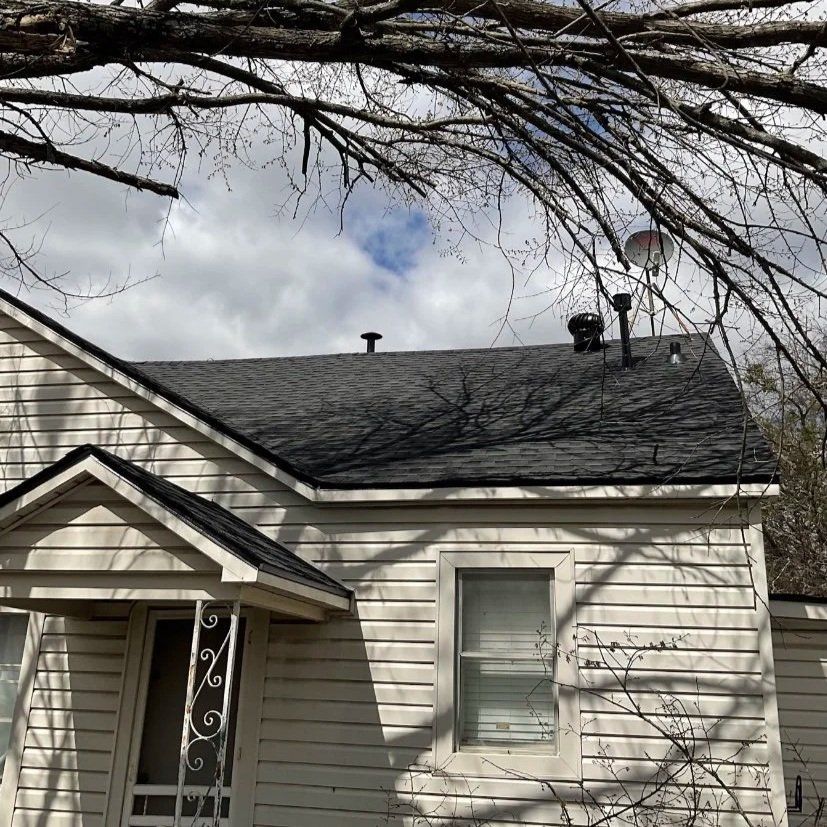 Small beige house with dark shingled roof under bare tree branches and cloudy sky.