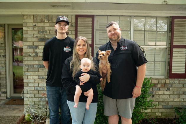 Family of four standing outside a brick house, holding a baby and a small dog.