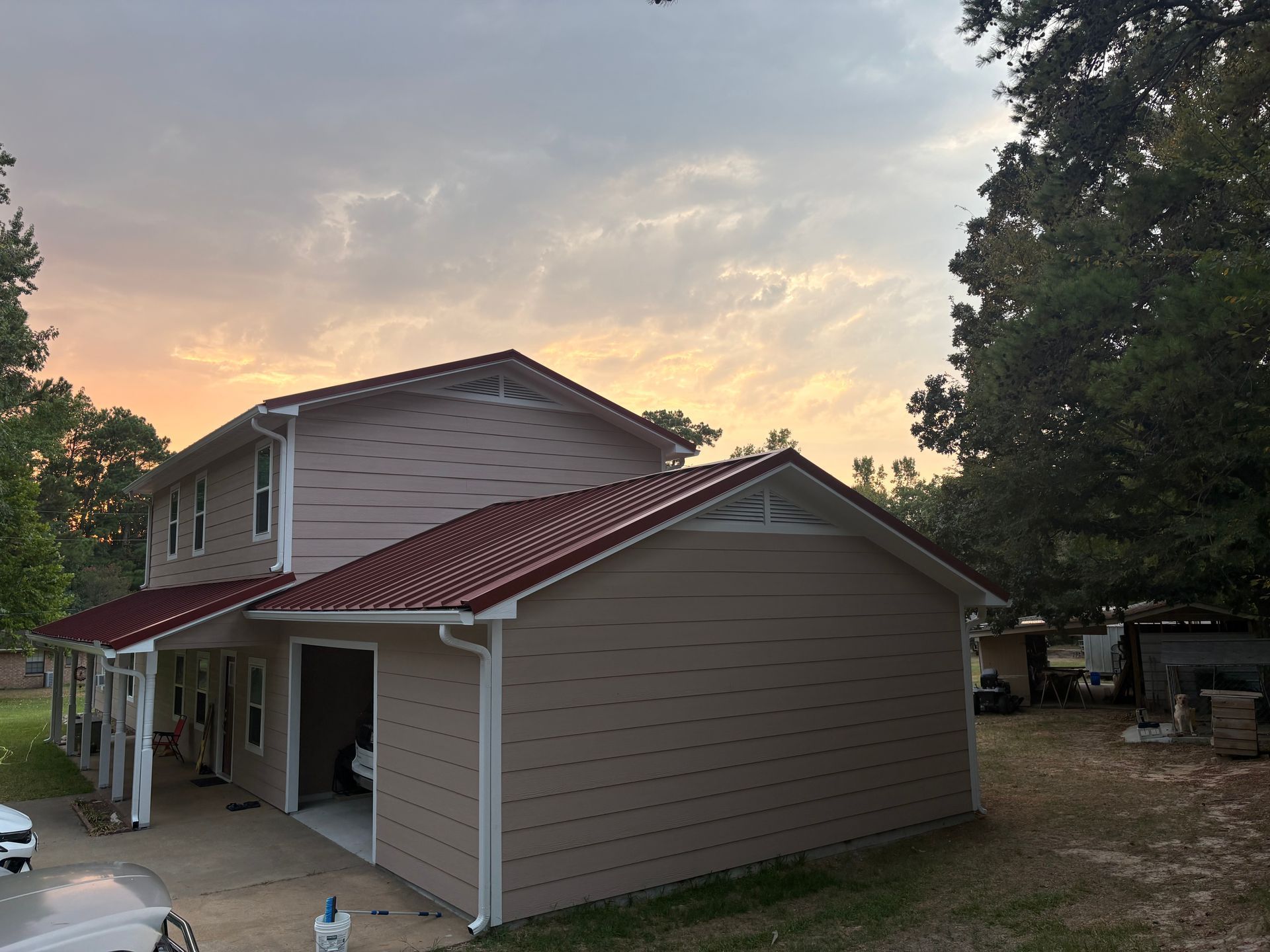 Two-story beige house with red metal roof and porch at sunset, surrounded by trees