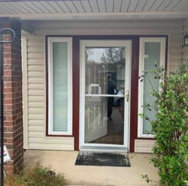 Front entry door with glass sidelights, maroon trim, brick wall, and a small porch mat.