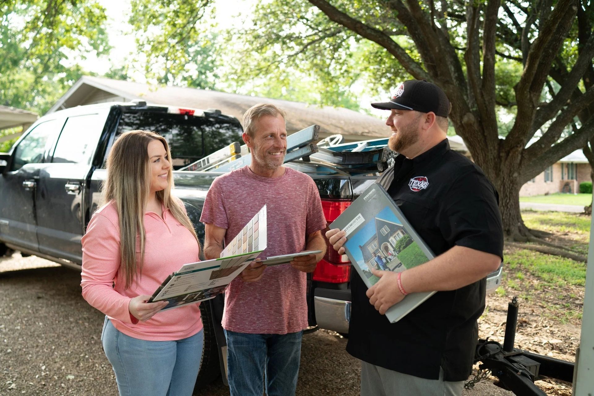 Three people stand by a pickup truck, smiling and holding items in a yard.