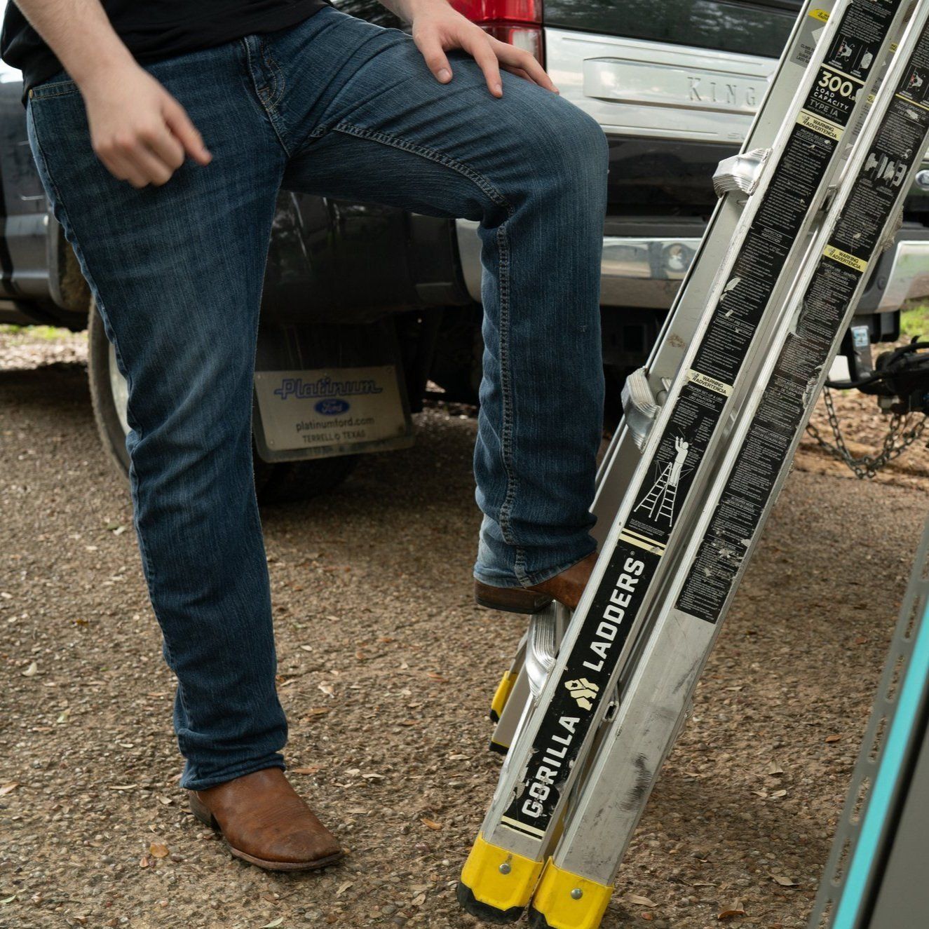 Person standing beside a ladder leaning against a vehicle, with a box and gravel ground nearby.