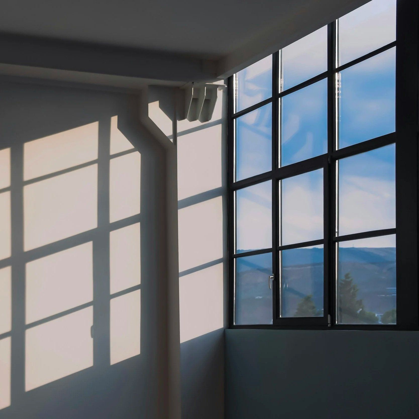 Sunlit room corner with large grid window, casting long shadows on pale walls and showing blue sky outside
