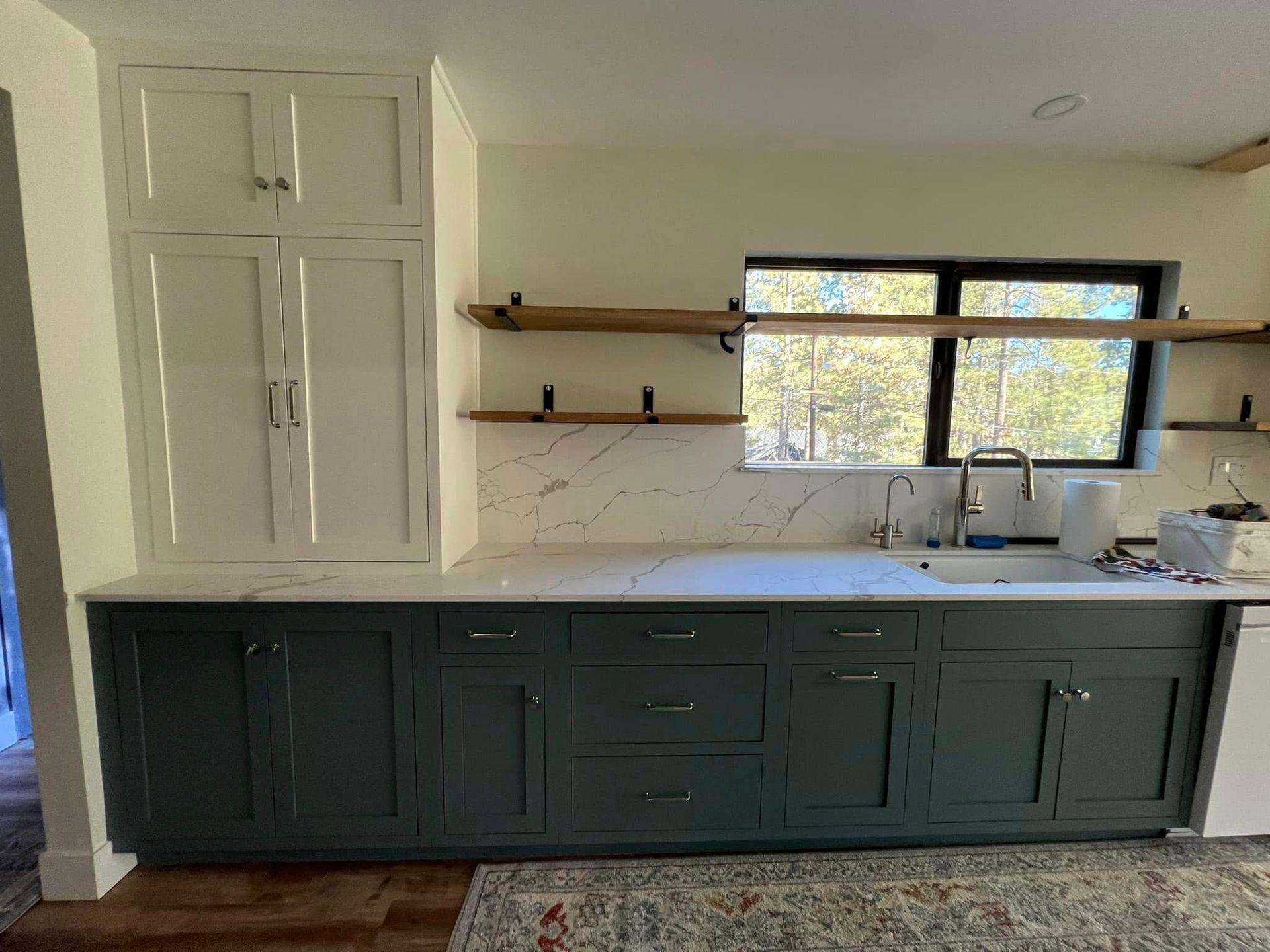 Green and white kitchen cabinets with marble backsplash and open shelving by a window.