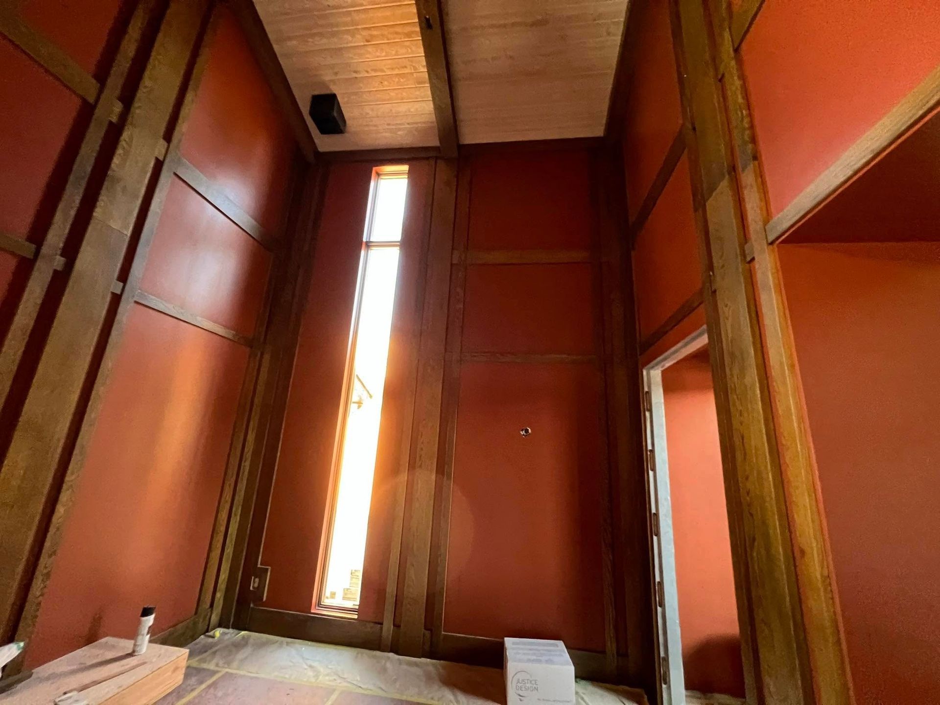 Interior of a room with tall narrow window, terracotta walls, wood beams, and an open door.