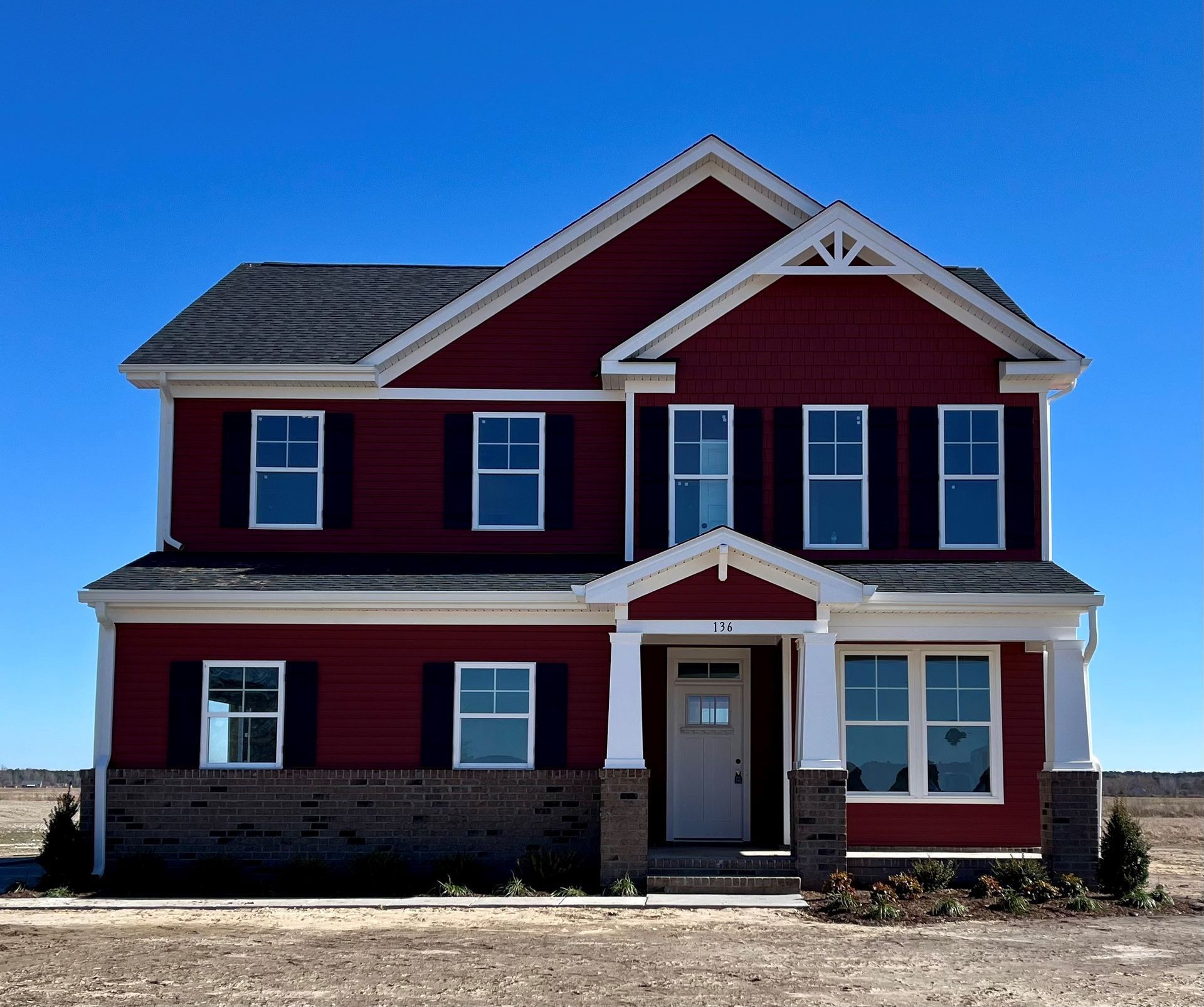 Two-story house with red siding, black shutters, white trim, and a brick base against a blue sky.