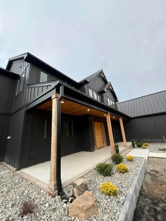 Black modern house with wood accents, porch, and gravel landscaping under a cloudy sky.