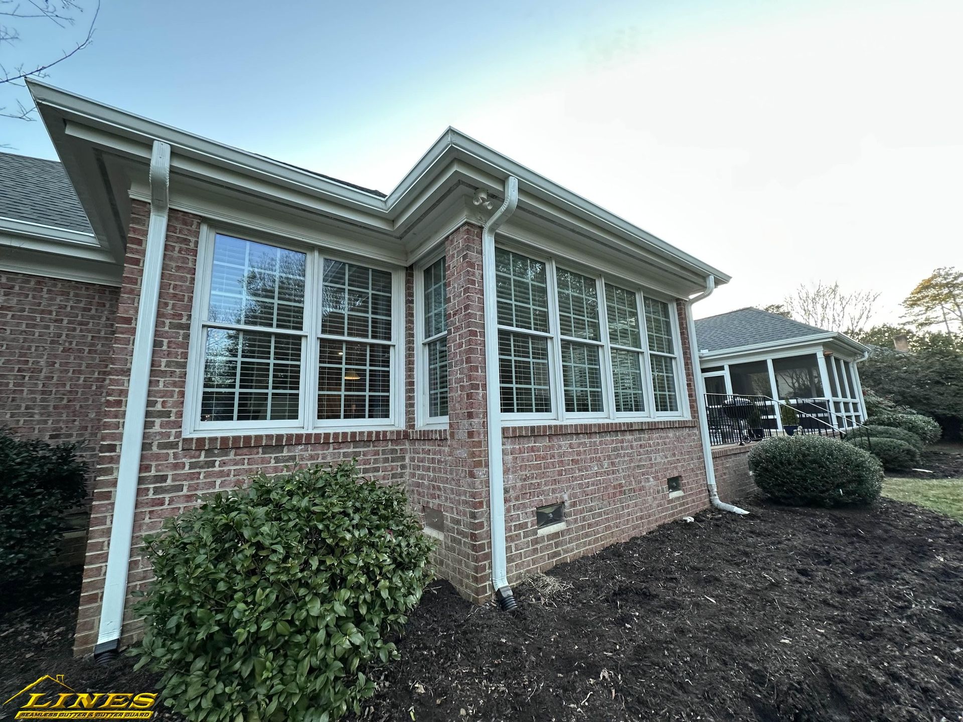 Brick house with white trim, gutters, and large windows. A green bush is in front.
