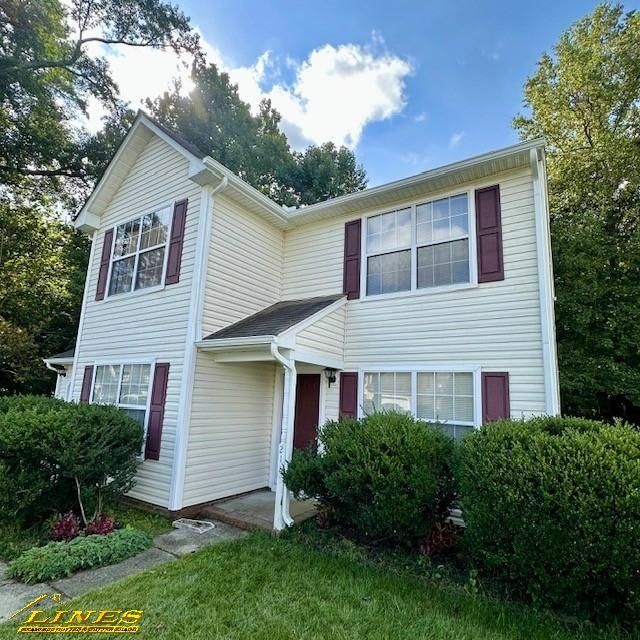 Two-story house with tan siding, burgundy shutters, and a small porch. Green bushes and grass in front.
