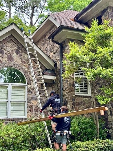 Two workers climb a ladder, carrying a long wooden beam, on a stone house. Green trees surround.