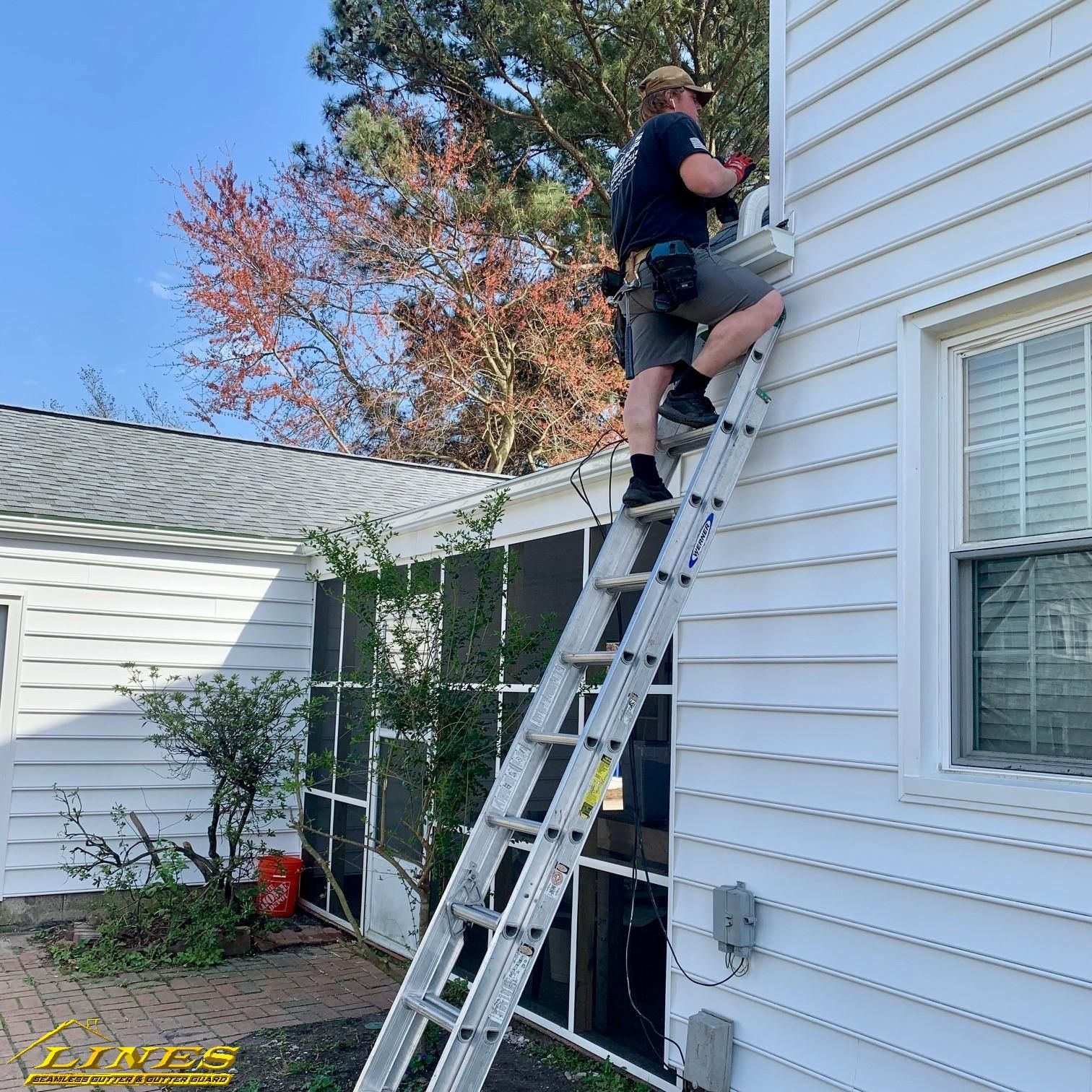 Man on ladder by a white building, working on gutters, holding tools.