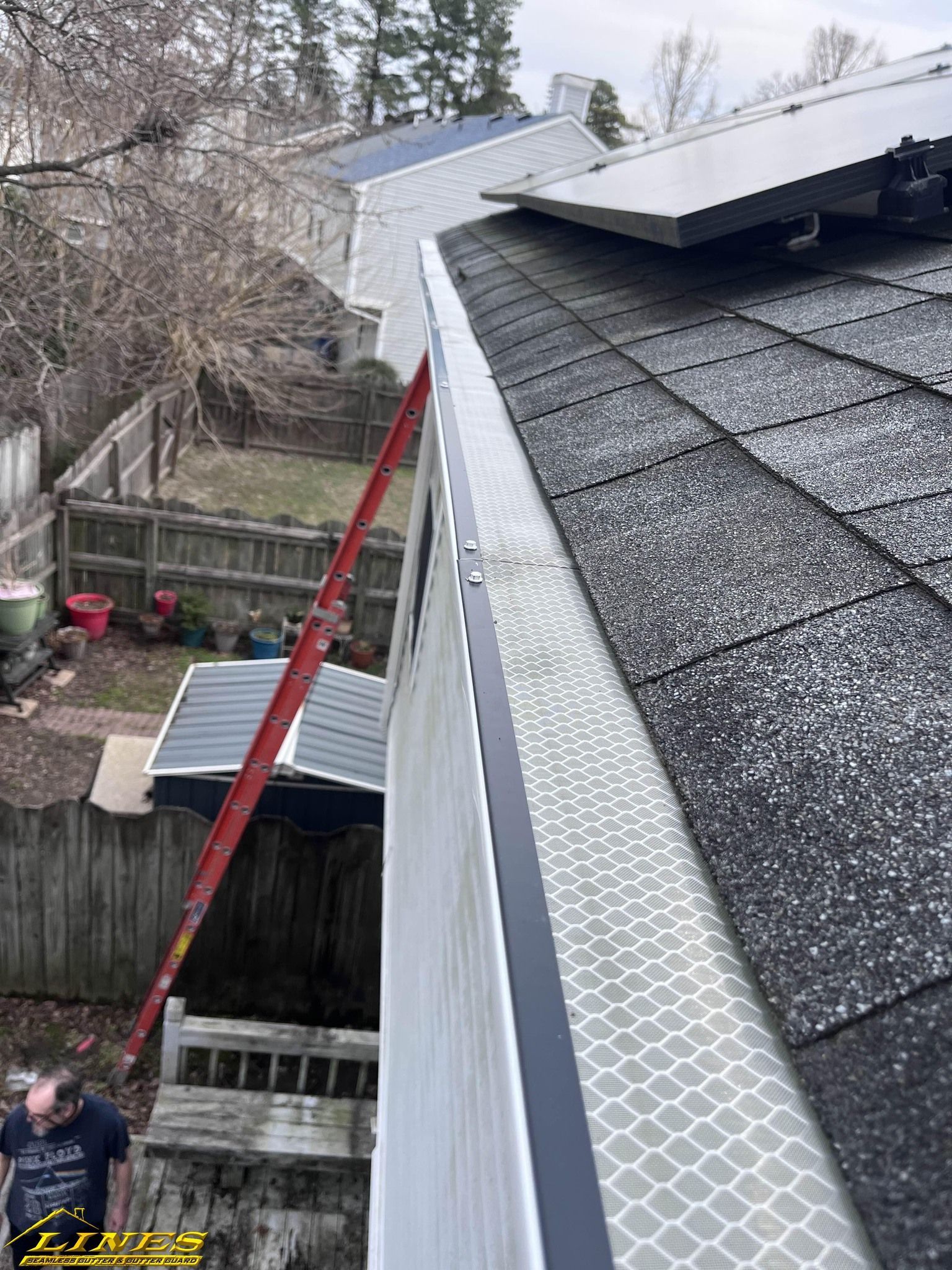 A person on a ladder working on a house roof with solar panels and a new gutter with a pattern.
