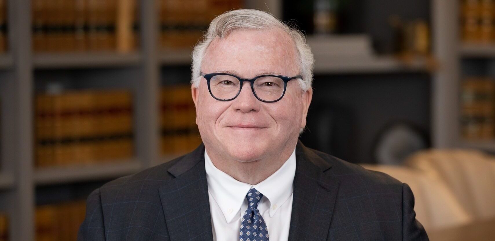 A man in a suit and tie is sitting in front of a bookshelf.