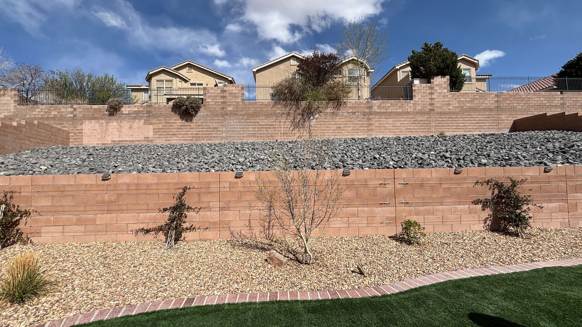 Terraced hillside with homes above, featuring brick walls, gravel, and sparse plants under a blue sky.