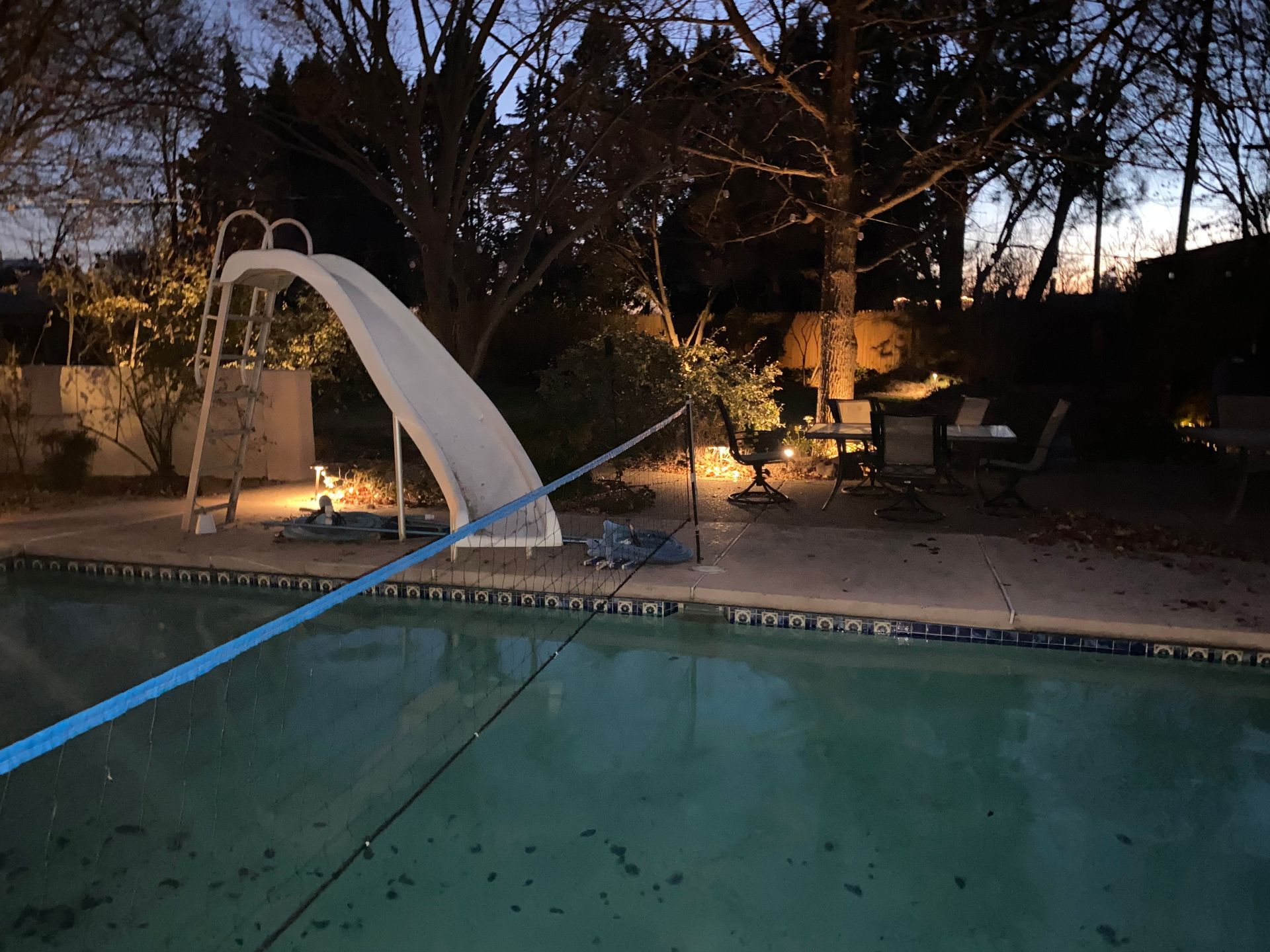 Backyard pool at dusk with a white slide, patio furniture, and trees.