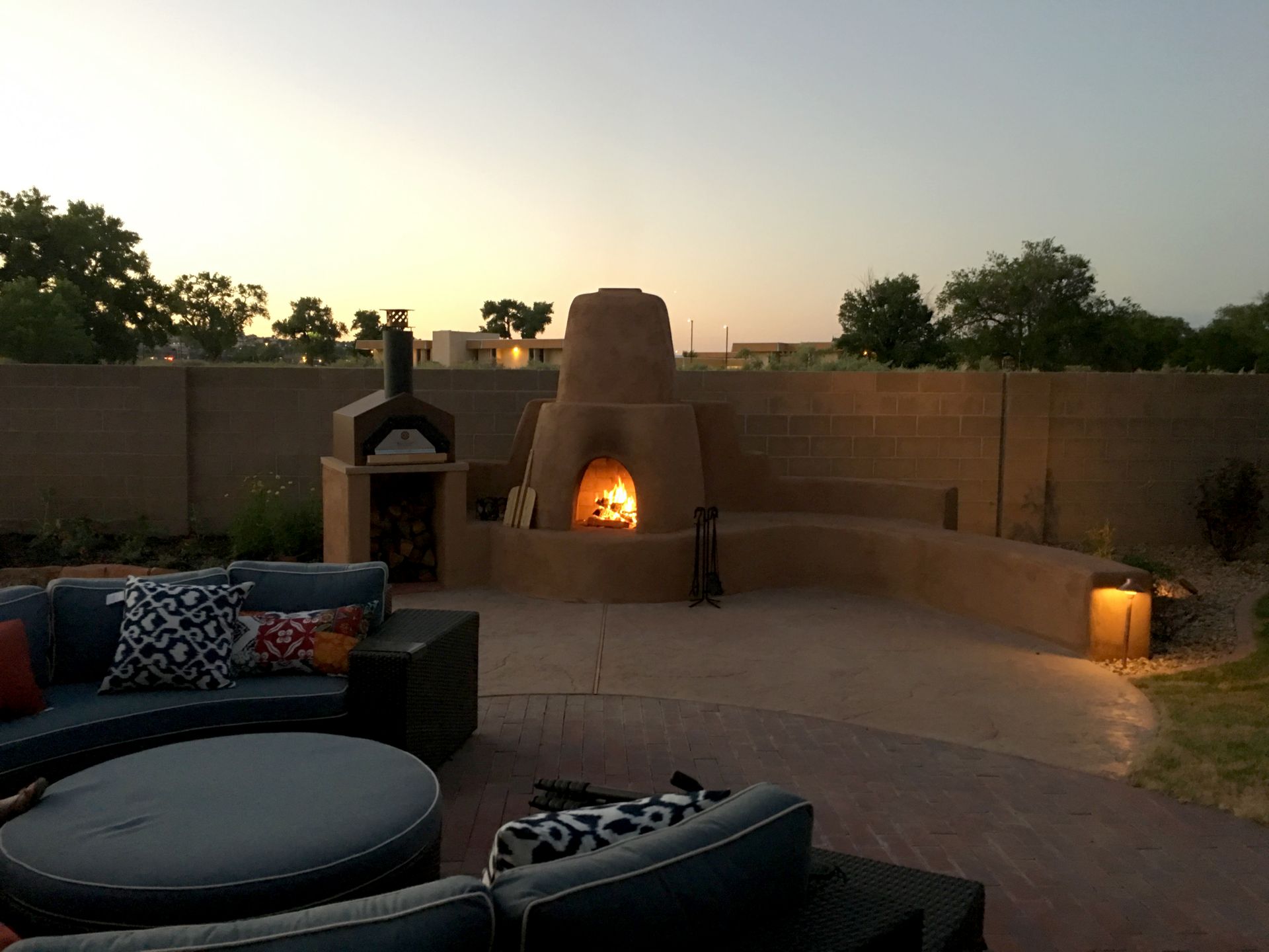 Backyard with a stone pizza oven, couch, fire burning at sunset.