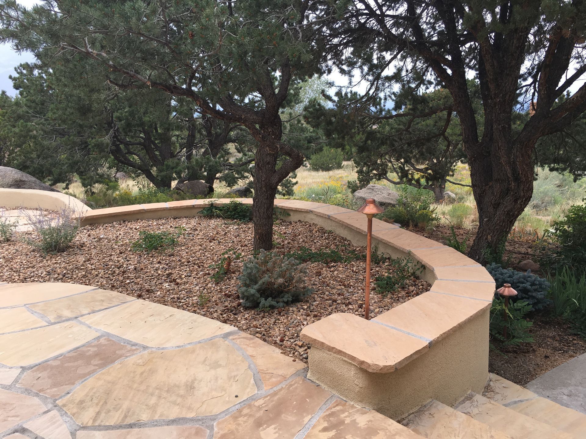 Stone patio with a curved retaining wall, trees, and a rocky garden in a natural setting.