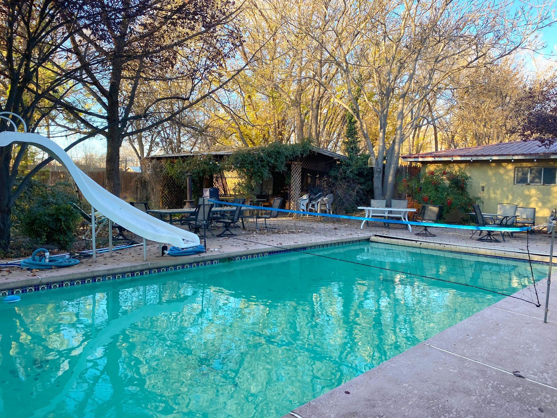 Pool with slide in backyard, surrounded by trees, tables, and a yellow building.
