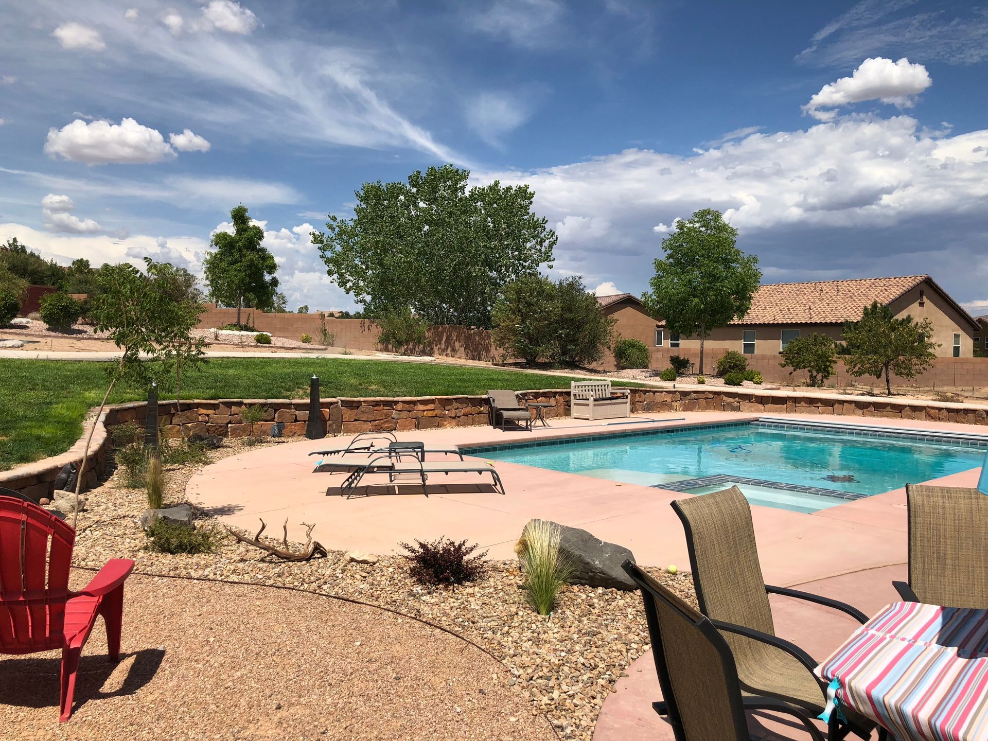 Backyard with pool, red chair, beige patio, and house under a blue sky.