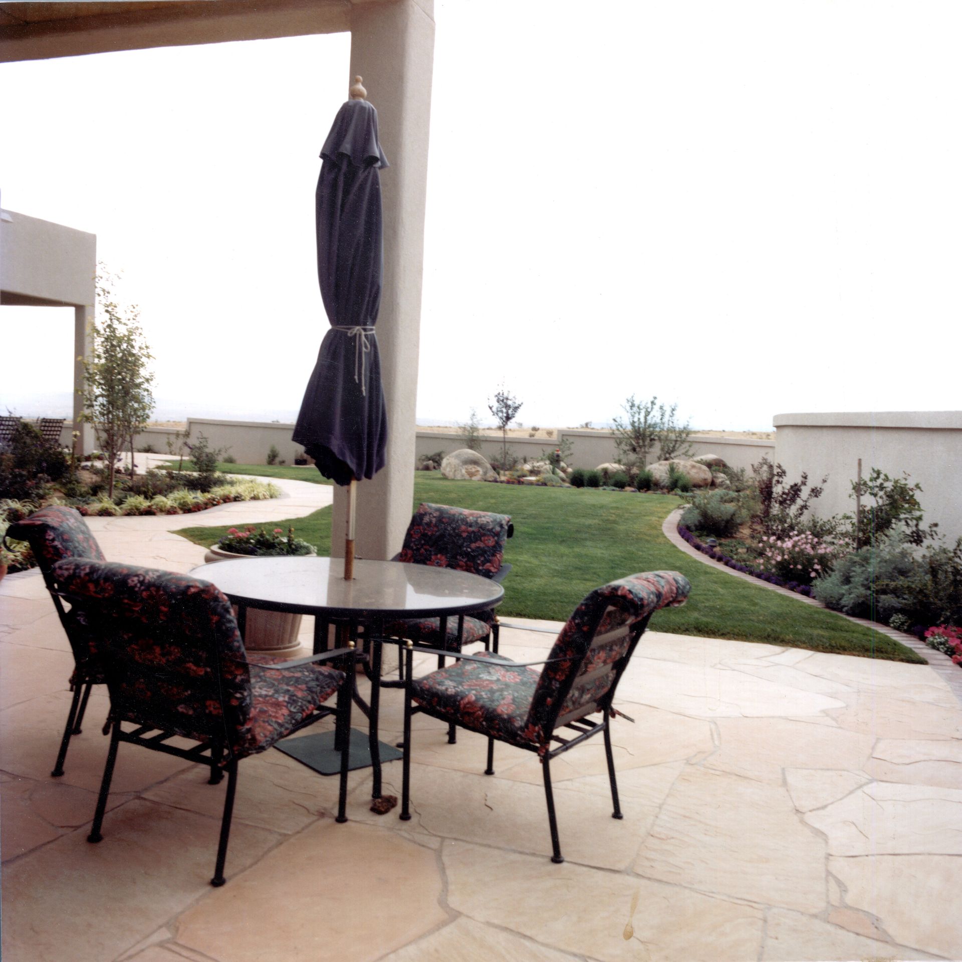 Patio with table, chairs, and umbrella; lawn and garden with a wall in the background.