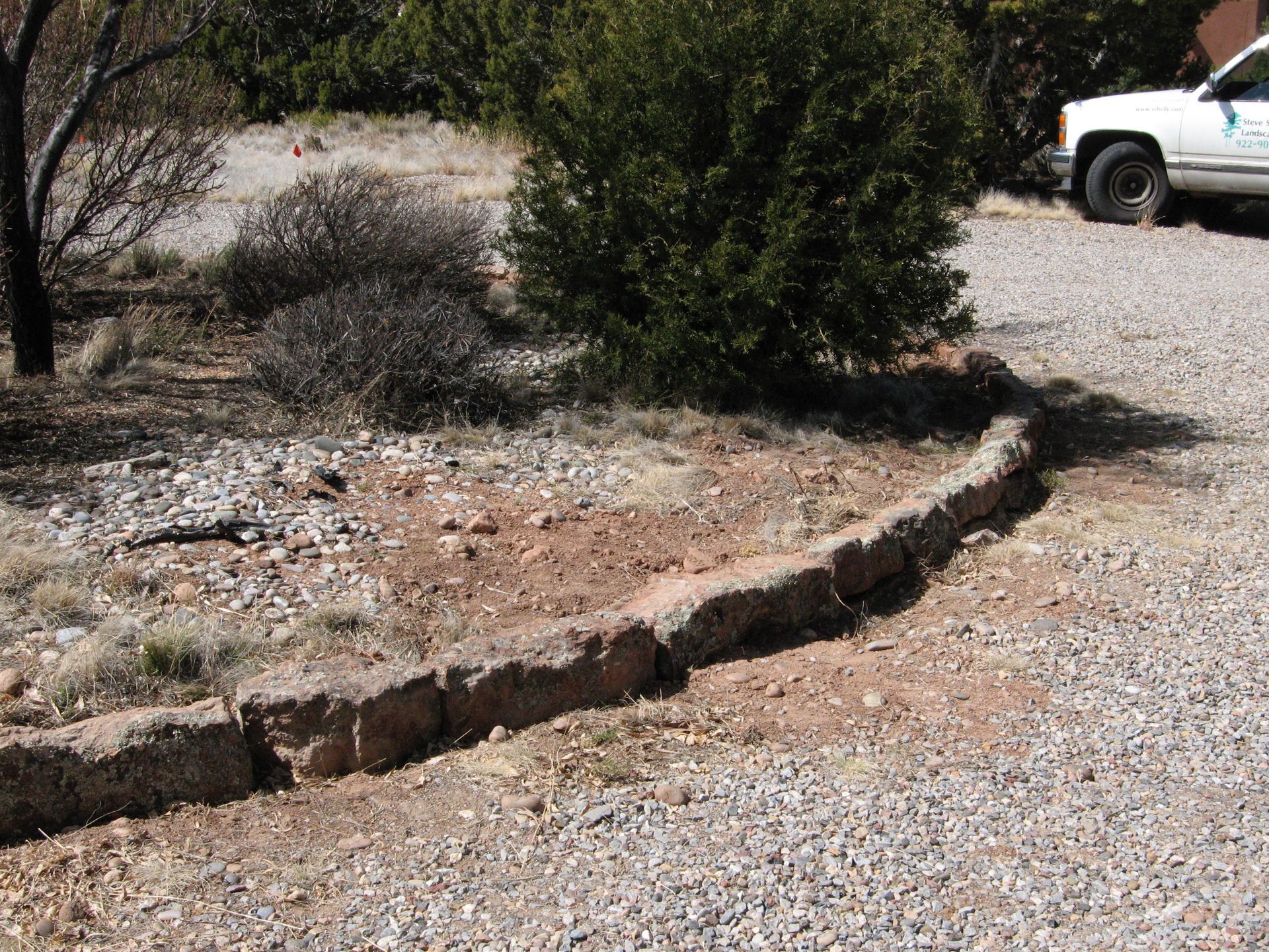 Stone border along a gravel driveway in a natural setting, next to a vehicle and shrubs.
