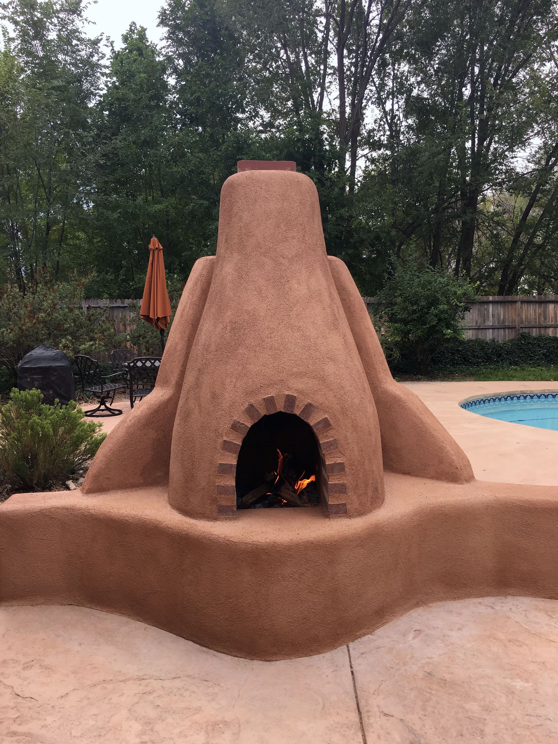 Adobe outdoor fireplace, with fire lit inside. Earth tones in a backyard setting near a pool.