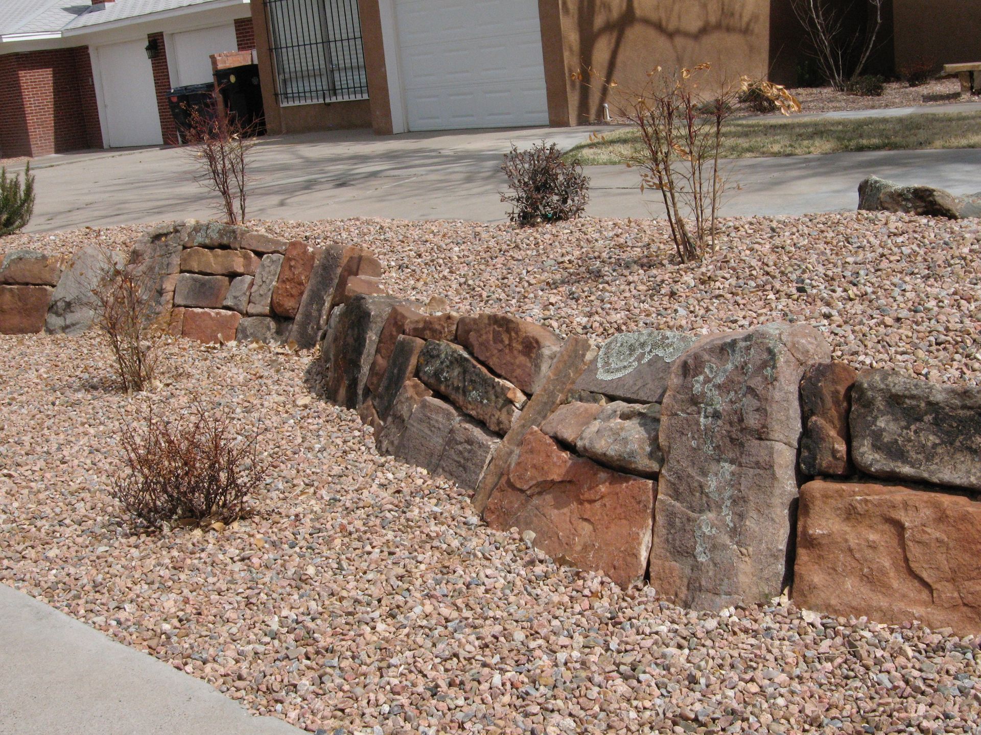 Low rock wall with sparse bushes in a yard covered in small, beige gravel.